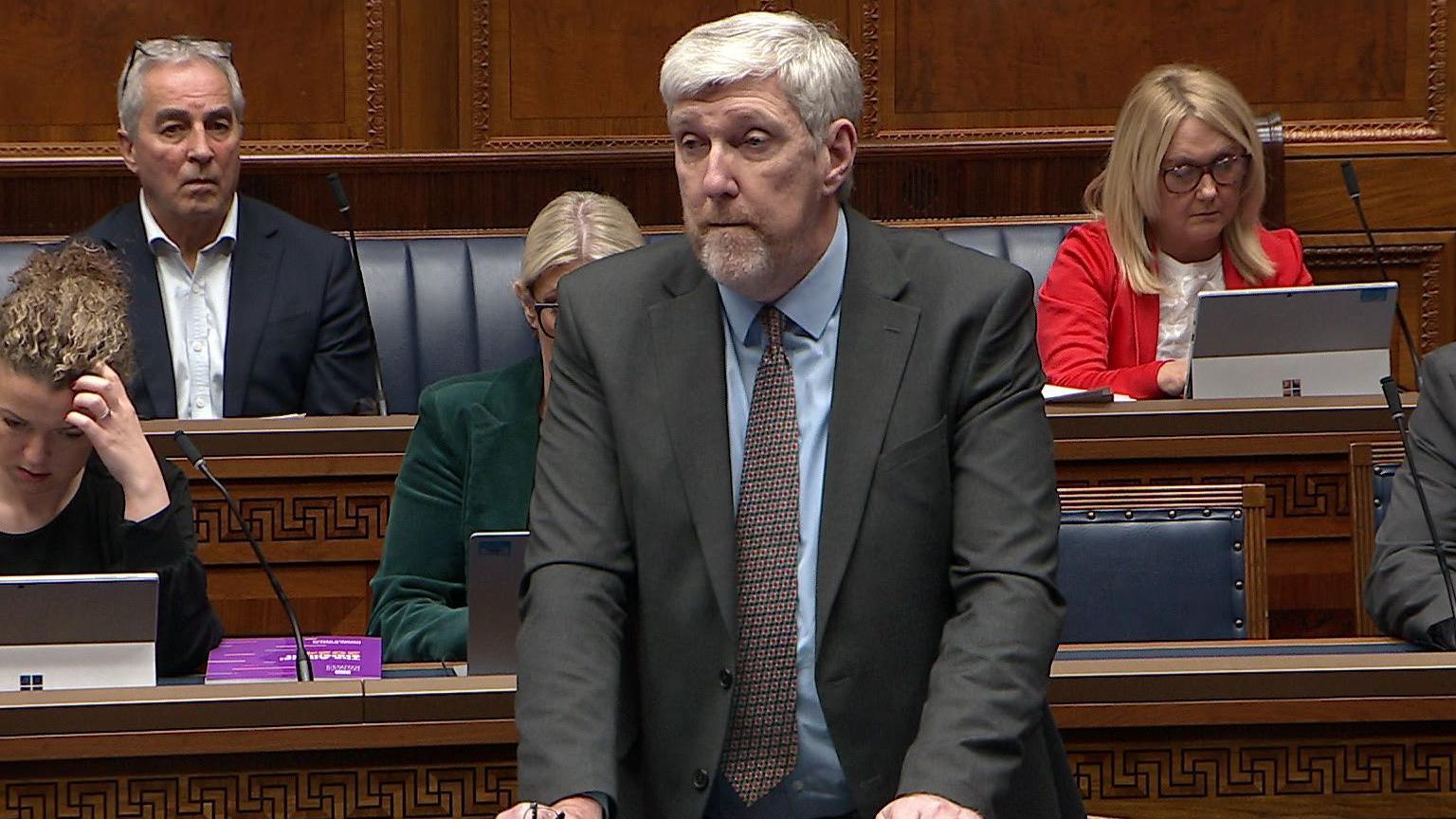 John O'Dowd wears a blue shirt, brown tie and grey suit. He speaks to MLAs in the Assembly chamber at Stormont. In his partially raised right hand, he holds a pair of black glasses. Behind him, sitting on the blue benches are three other MLAs.