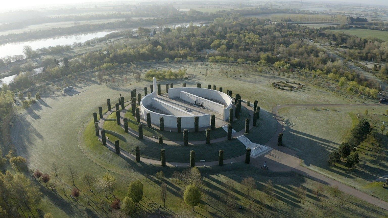 An aerial view of a memorial at the National Memorial Arboretum. There are four circular levels. There are tall bushes decorating each layer as well as brown pathways. On the top level is a circular concrete structure split into two semi-circles.