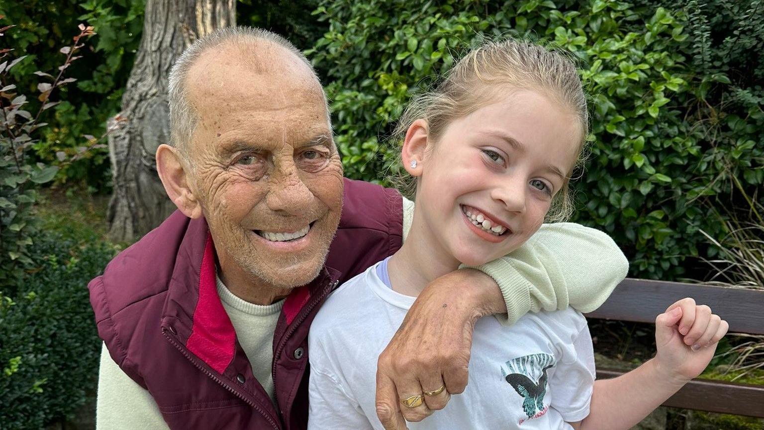 Dave on the left wearing a light-green jumper with a burgundy-coloured gillet. He has his arm around his granddaughter Abbie, who is on the right. She has blonde hair and is wearing a white t-shirt