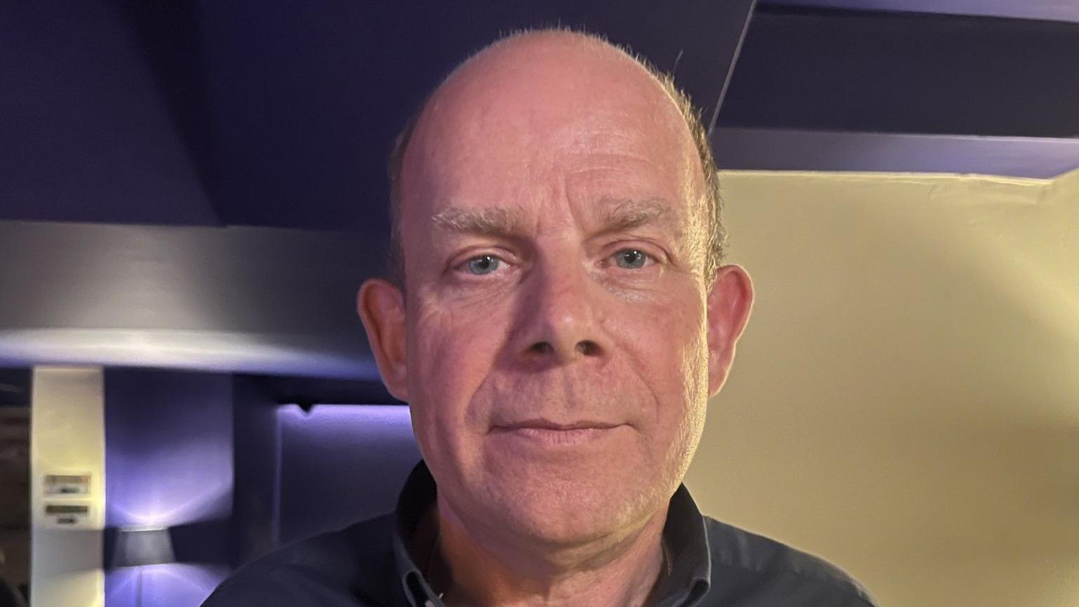 A head shot of Philip Romeril. He is wearing a black shirt and he has receding hair and blue eyes.