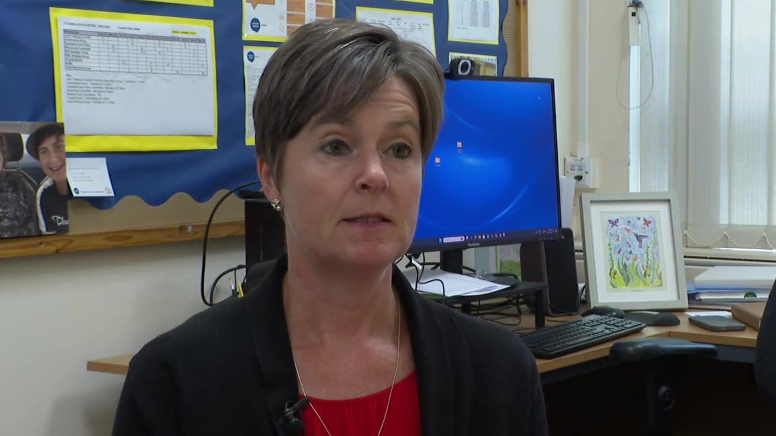 Sue Julyan wearing a black blazer, red top underneath and silver necklace sat in front of a computer on a desk which also has a colourful piece of artwork on it. Behind her on a board is a photo of a smiling young man, possibly a teenage relative of hers. Sue has a short haircut with dark hair tinged with grey.