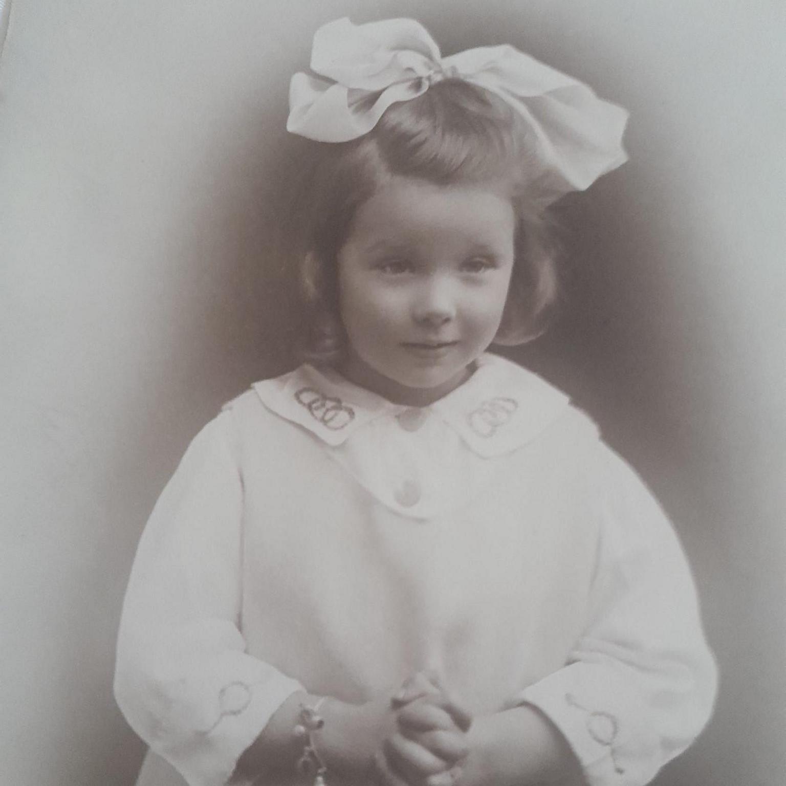 An old black and white picture of a girl with her hair tied up in a large white bow and wearing a white gown.