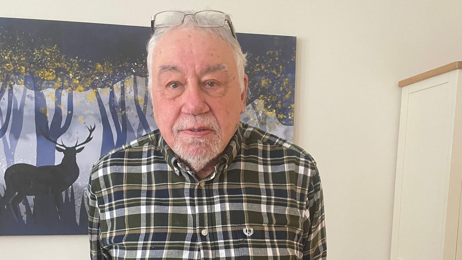 Geoffrey wearing a green, white and yellow checked shirt, glasses balanced on top of his head, sporting a closely cropped goatee and standing in front of a picture of a stag in a forest. 