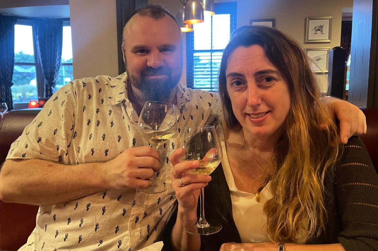 Mark Willerton with an arm around his partner Lynne. It looks like they're at a restaurant or gastropub. They are smiling while holding up glasses of white wine.