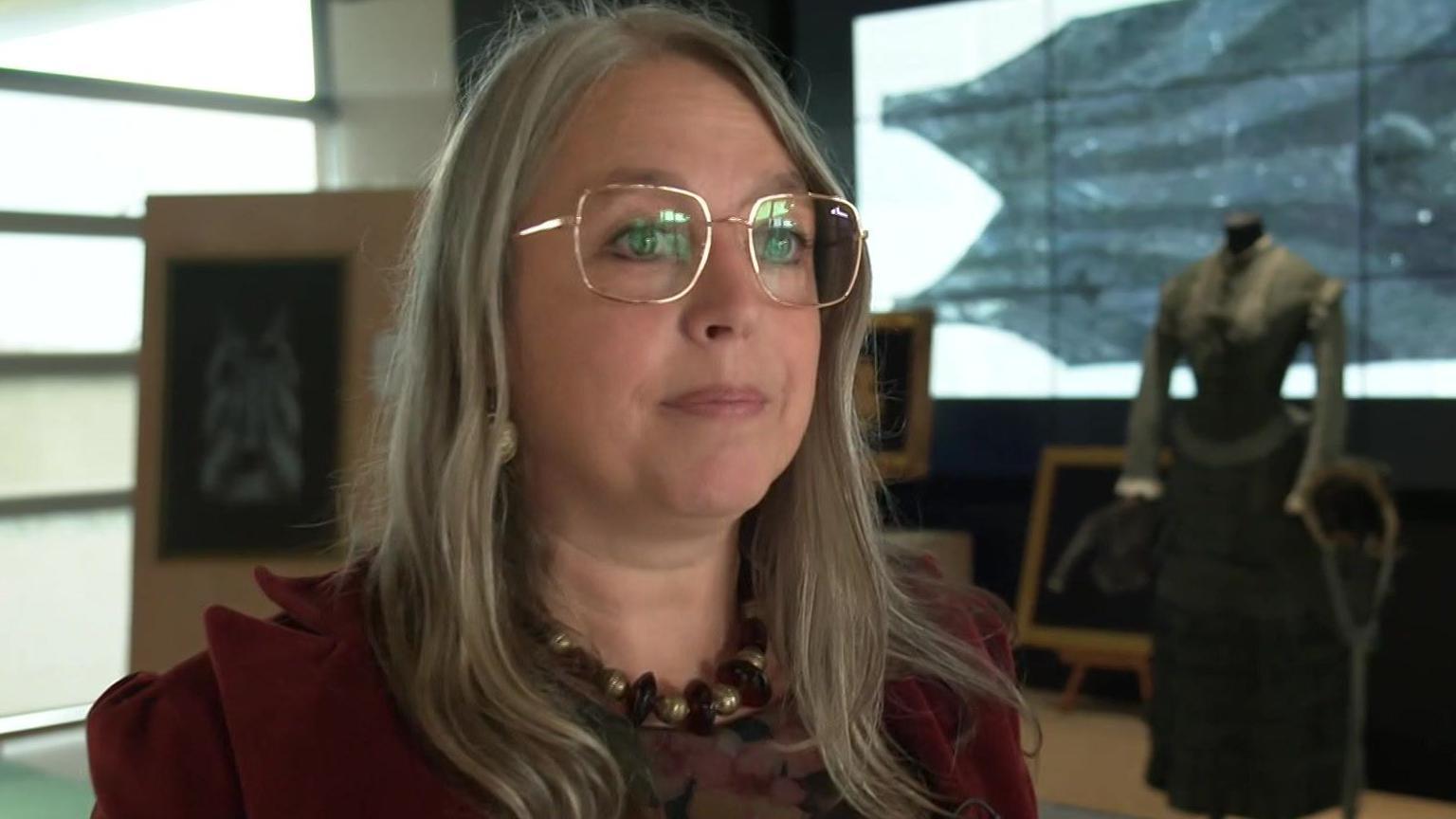Sian Pilley looking to the right. She is wearing gold-framed glasses and has blonde hair. She is wearing a dark red velvet jacket and a necklace with large gold and dark red beads. To her right can be seen one of the dresses in the exhibition.