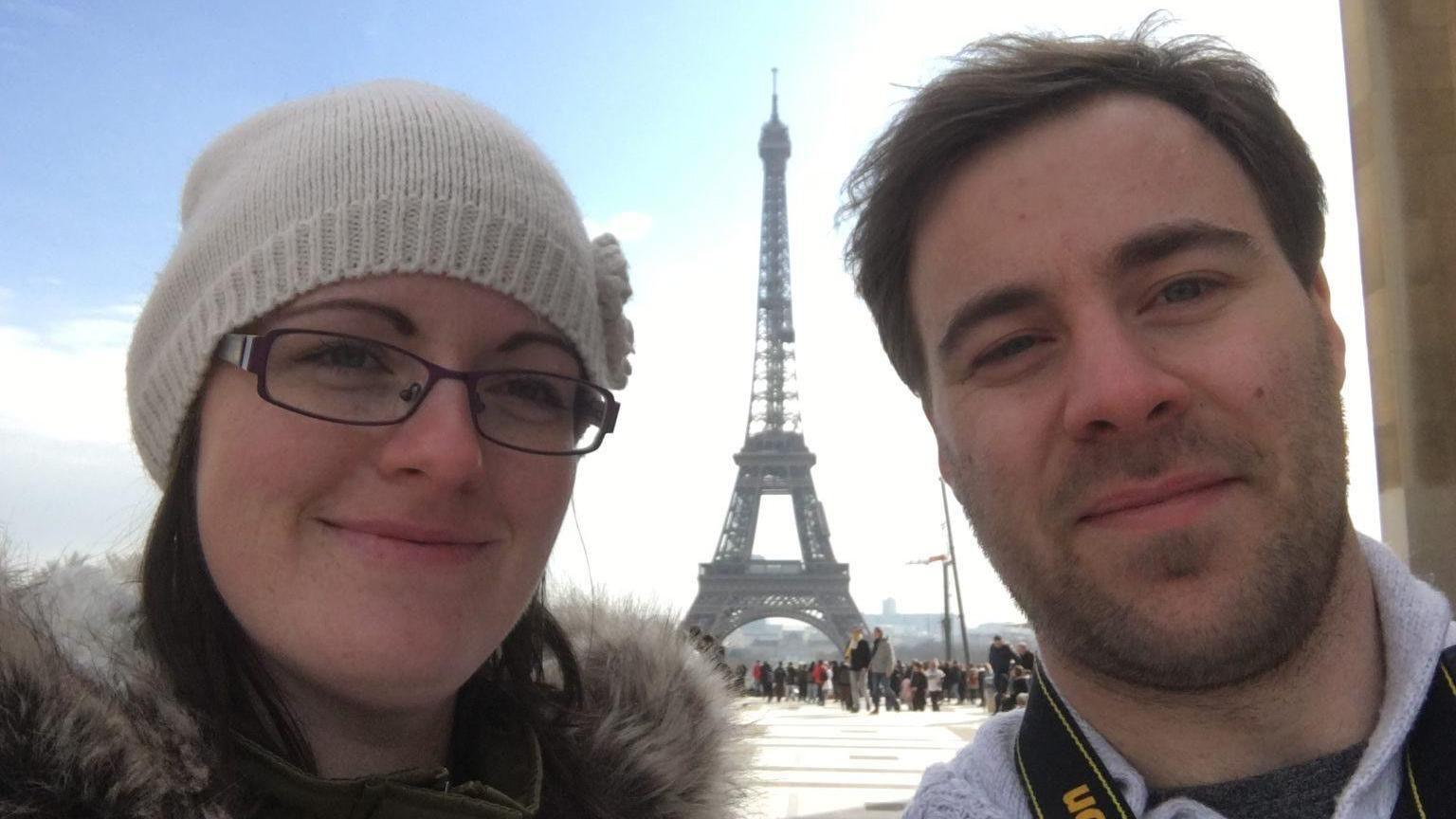 A close-up image of a woman and a man, Nikki and Sam Perett, with the Eiffel Tower in Paris in the background. Ms Perret has brown hear and is wearing glasses and a white beanie hat. Mr Perret has short brown hair and is wearing a white top with a collar.