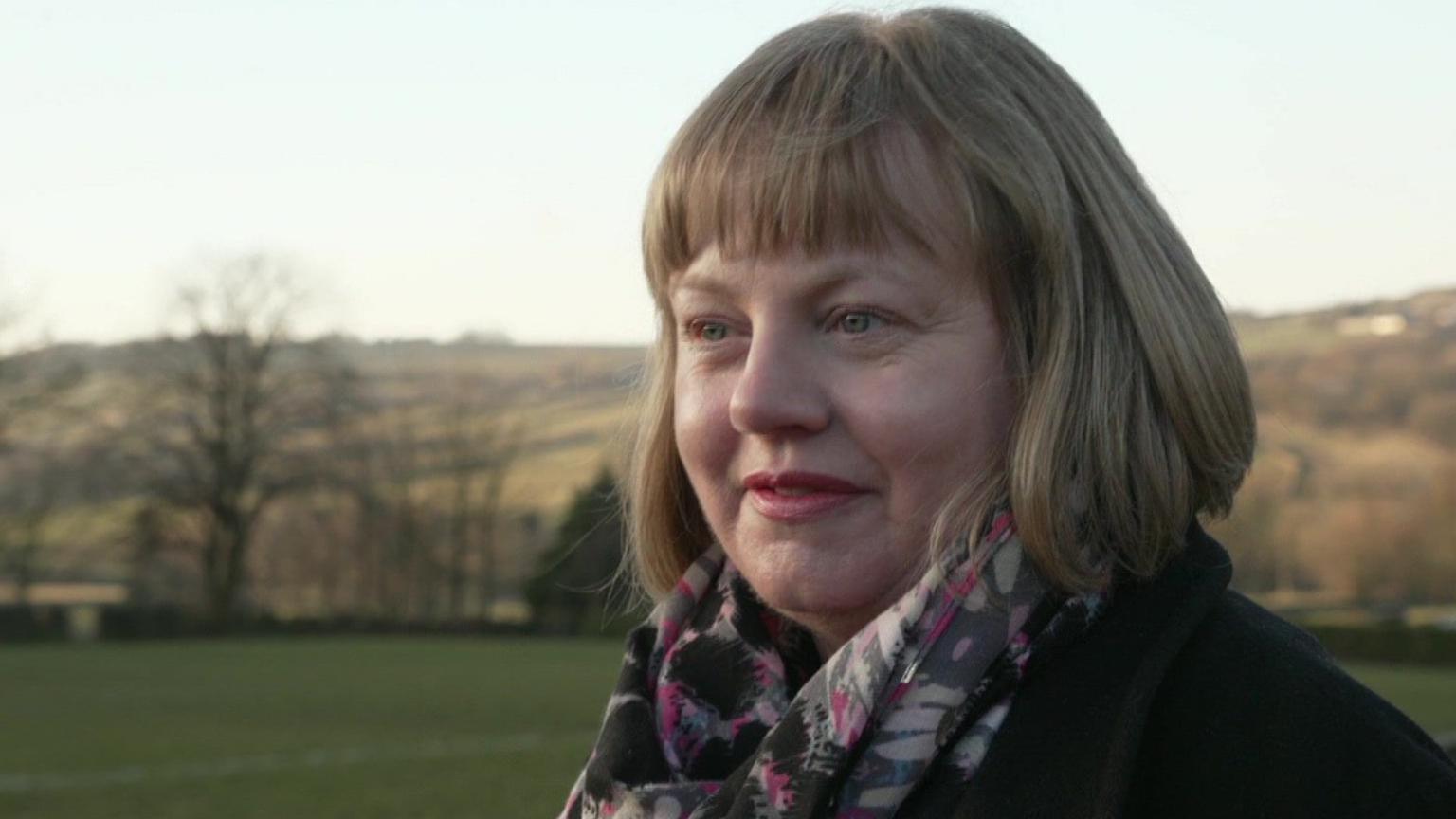 A woman with dark blonde bobbed hair stands in front of a rural backdrop. She wears a coat and scarf.