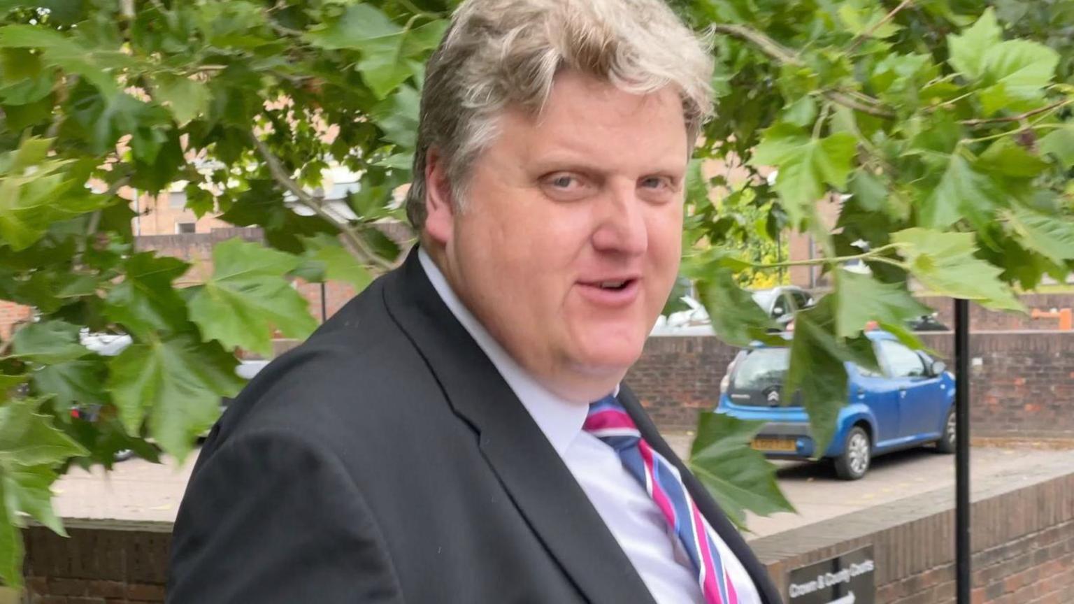 Gregory Hill, a man in a black suit jacket, white shirt and striped tie. He has blonde hair and is pictured outside court in Norwich. A tree, a wall and a blue Citroen car are behind him.