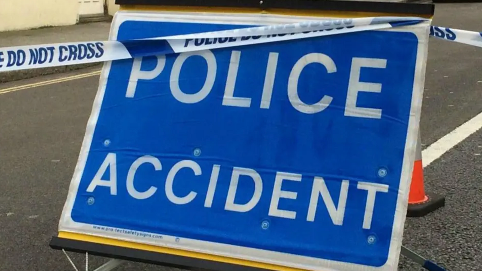 A blue police sign that says 'Police Accident' in white writing. It has been placed on a road.There is police tape going over the sign and an obscured orange cone behind it.