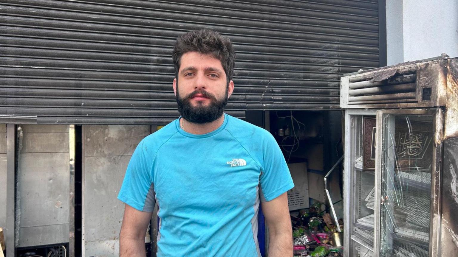A bearded man looking into the camera, from outside a burnt-out shop with its shutters half down. A smoke-damaged fridge and piles of damaged goods are visible behind him.