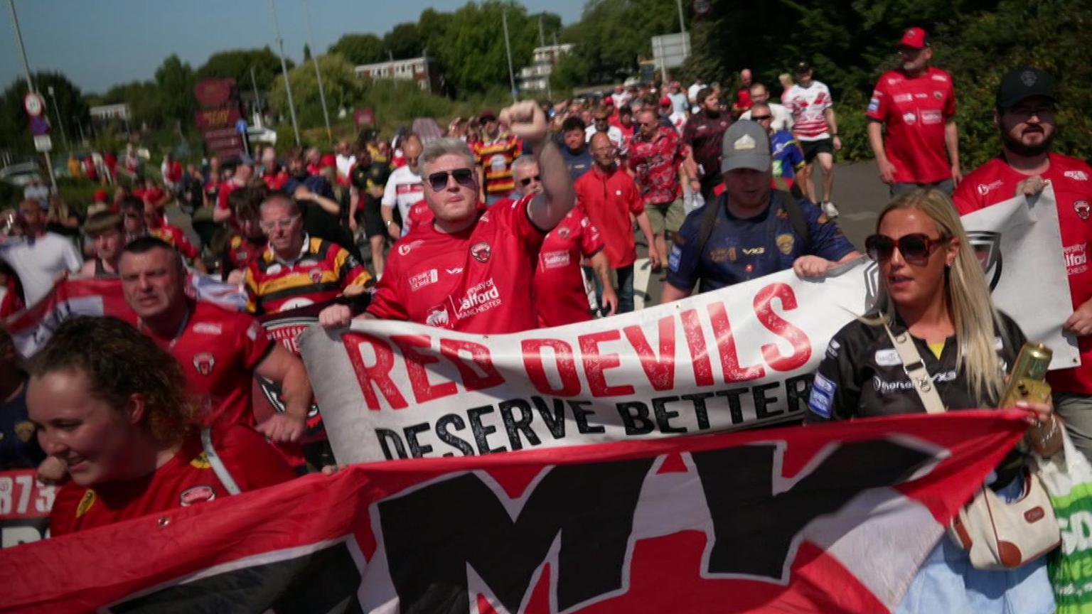 A crowd of Salford Red Devils fans march together, dressed in the club's red and black colours. One banner reads "Red Devils deserve better".