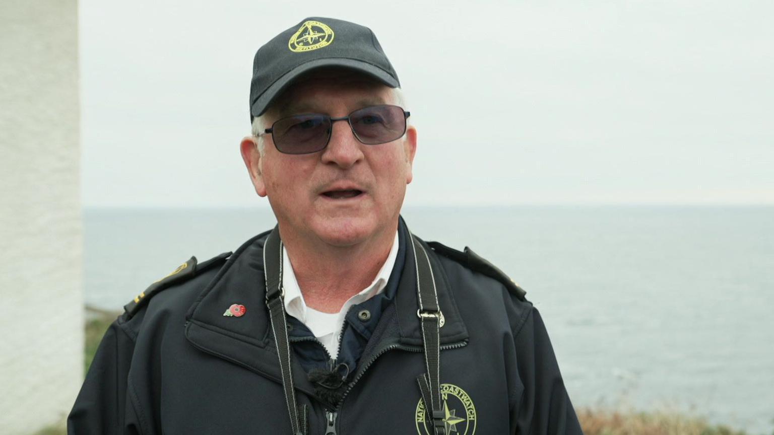 Jerry Betteridge stands in front of the coastline. There is a grey November sea behind him and the edge of a white wall of the lookout post to his side. He has a camera strap around his neck.