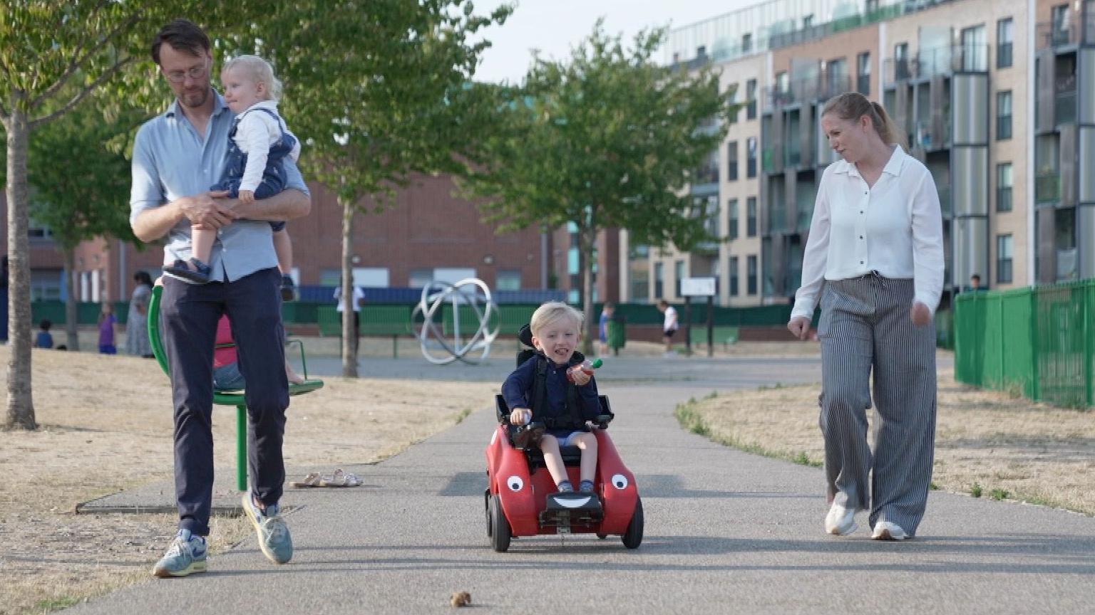 A young family - mum, dad and two boys, one a toddler in his dad's arms and the other a four year old in a motorised toy vehicle - walk together in a grassy area near blocks of flats. 