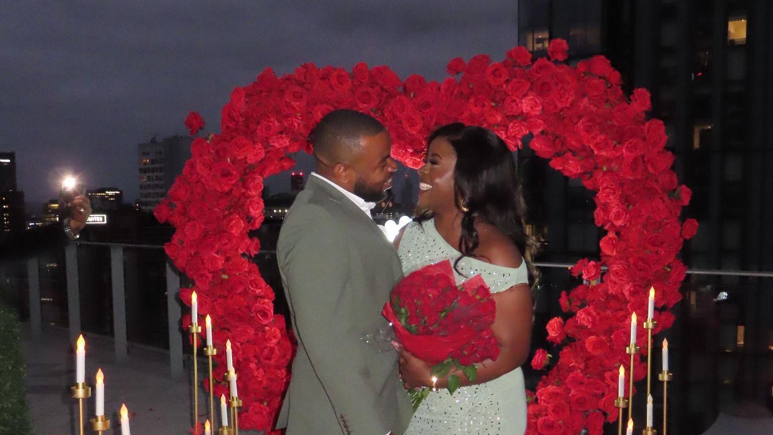 Side profile of Fred and Malaika facing each other as they look into each others' eyes in front of a large heart-shaped stand of red roses with candle lights on both sides. It is night and high-rise buildings can be seen in the background behind the veranda.
