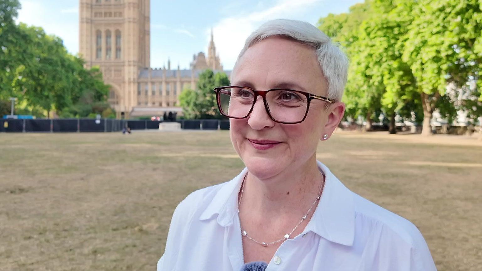Woman with short white hair, glasses in a white top stands in Westminster park looking into the distance.