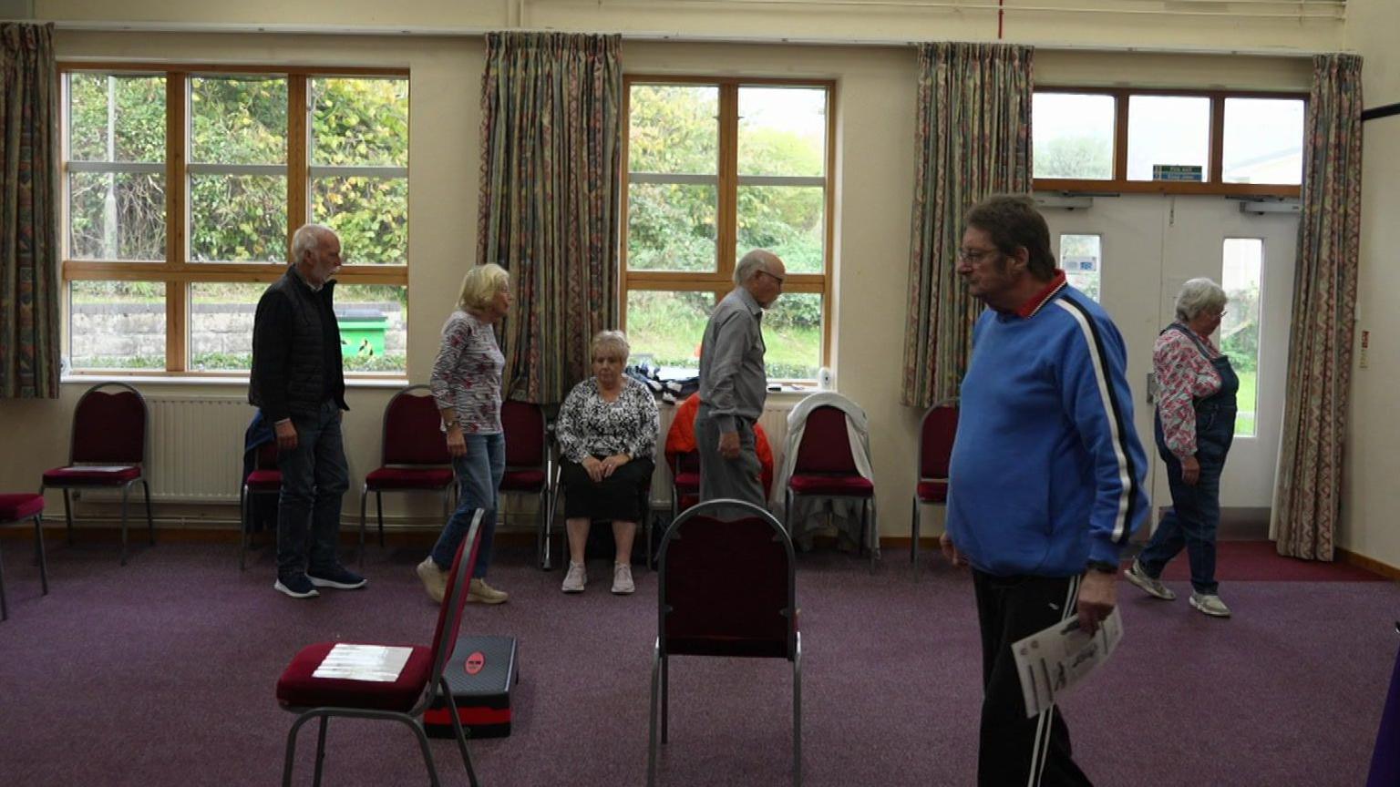 Six people walk around a room practicing breathing exercises. The walls are magnolia and the carpet is deep purple. 