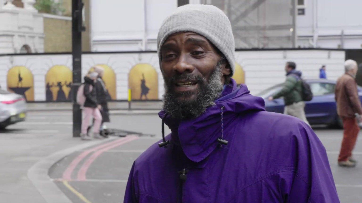 Tony, on a busy street in Victoria. He wears a purple coat and grey beanie hat.