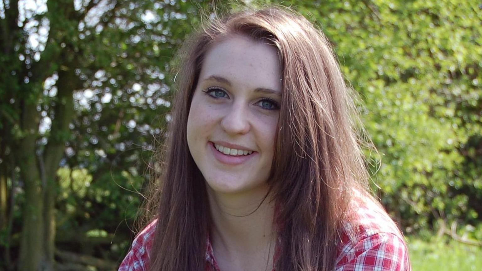 Alice Ruggles smiles while sitting on grass. She has long brown hair and is wearing a red-checked top.