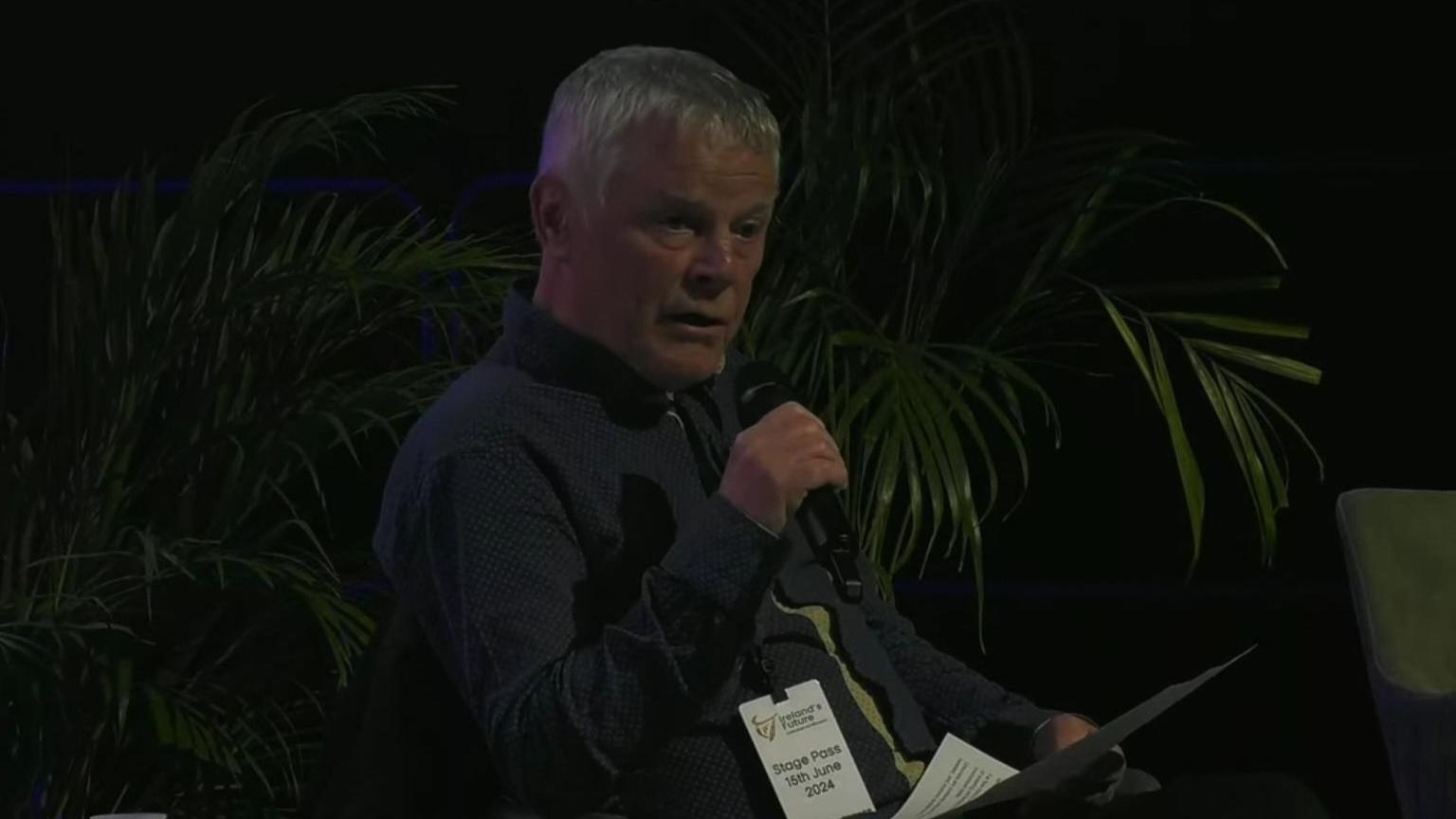 White haired man sitting and speaking at event