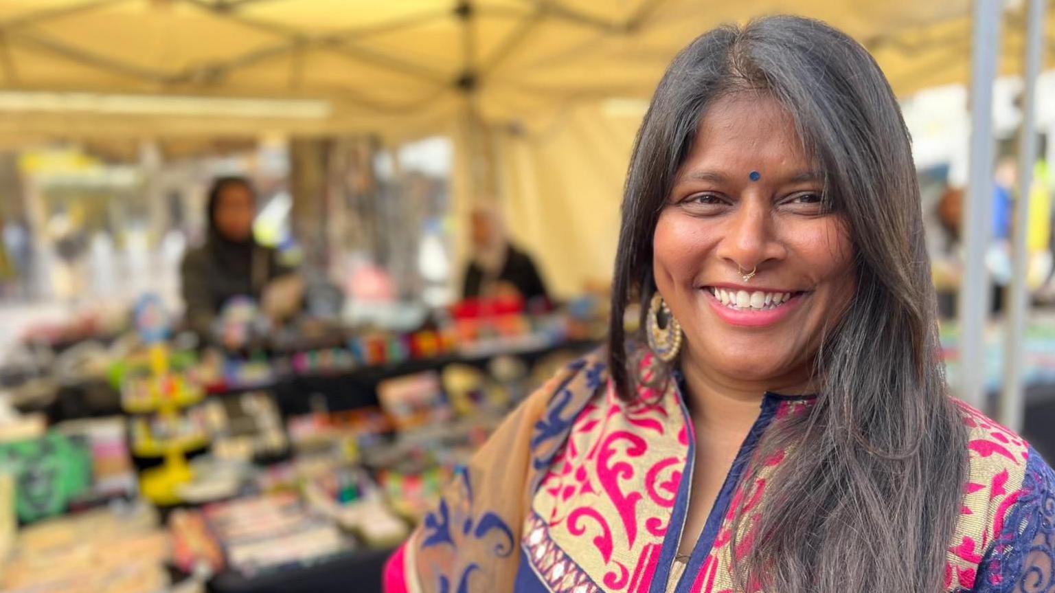 Leena Patel who has long black hair swept over one shoulder. She is wearing a top with a purple body and its shoulders and arms have pink white yellow and purple lace patterns. She is smiling towards the camera and behind her are stalls with out of focus goods on them within a tent