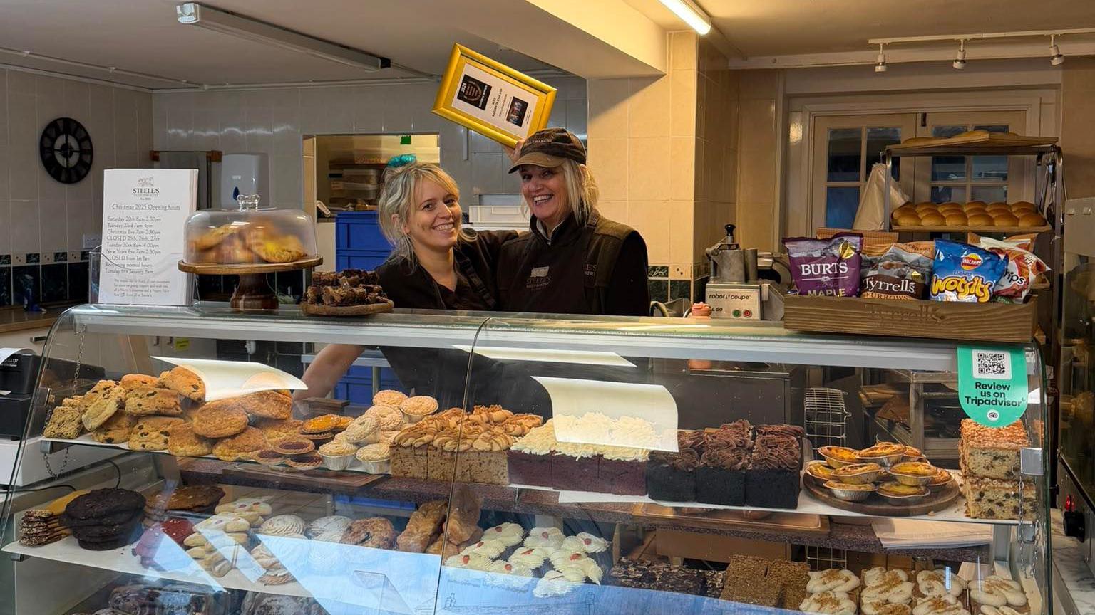Two blonde women behind a glass bakery counter with stacks of cakes, pies and baked goods behind it. One of the women is holding up a gold-framed photo with an award inside it