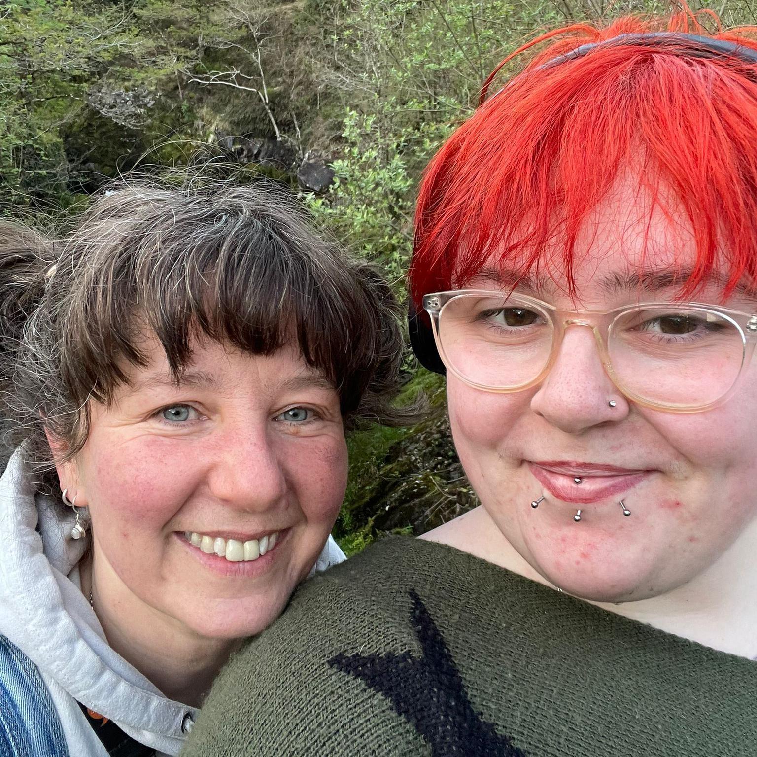 Elliw a'i ffrind yn tynnu selfie. Mae ganddi wallt coch llachar. Mae goedwig yn y cefndir. 