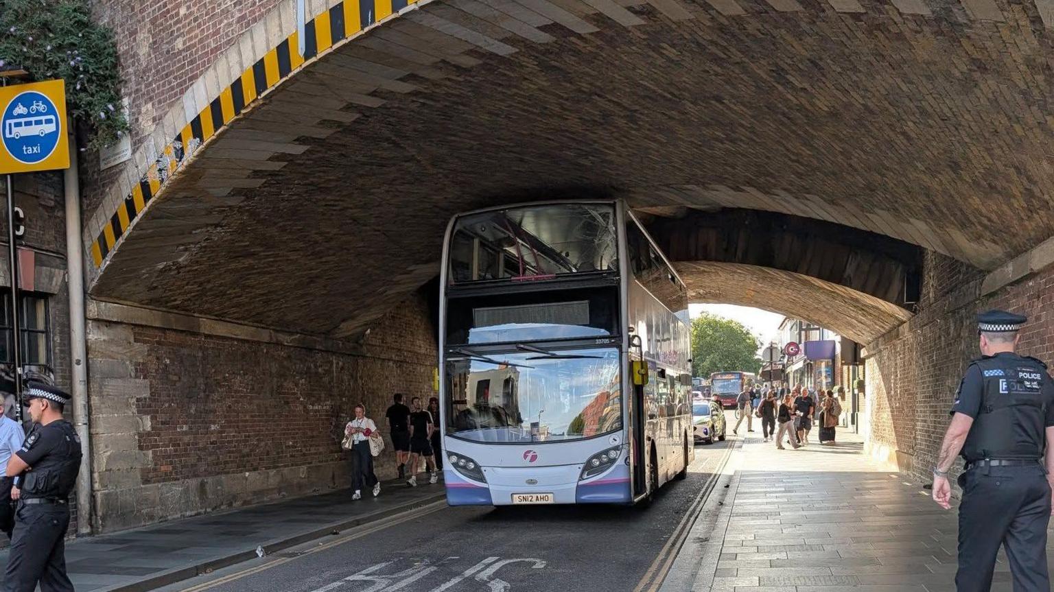 A blue double-decker bus going under a low tunnel, a police officer is on the path to the right