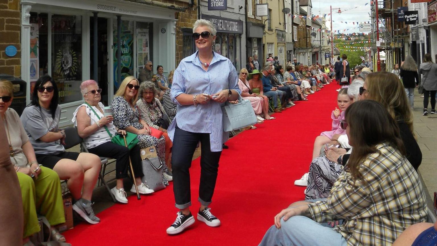 A woman with short white hair and dark glasses poses on a red carpet in a shopping street, wearing a blue shirt, light-coloured handbag, denim trousers and blue and white plimsolls. There are spectators sitting on both sides of the carpet, and shops behind them.