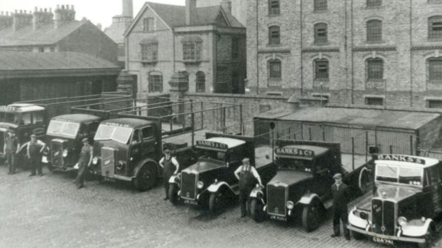 A black and white photo of a row of lorries with people in suits and hats standing in front of them and a large brick building with several floors in the background