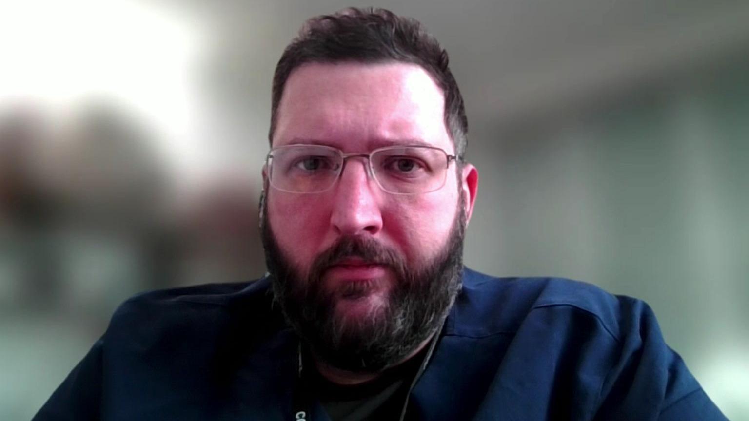 A man looks into the camera. He has short brown hair and a beard and wears glasses and is wearing a dark blue shirt. The background is blurred.