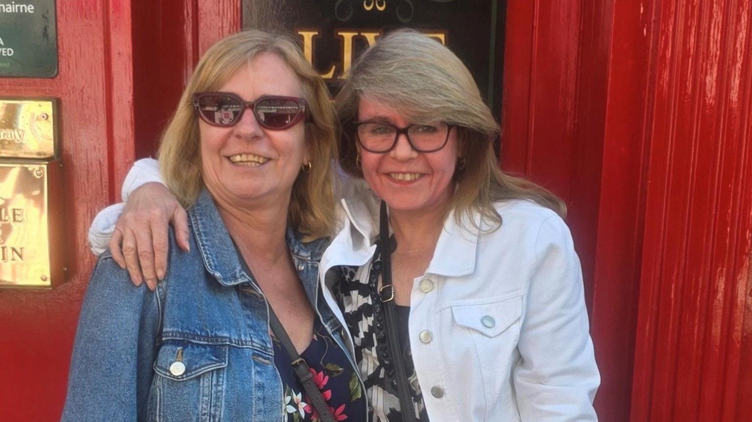 Yvette Greenley stands with her arm around her sister Beverley in front of a red door. Both are wearing denim jackets.