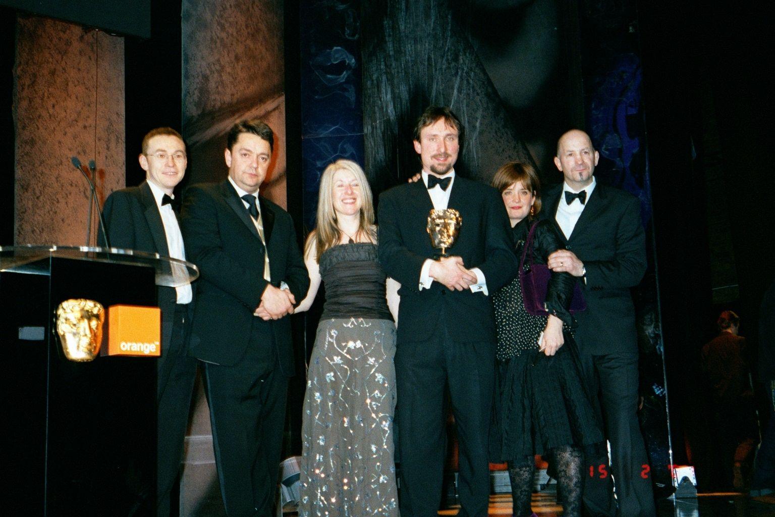 Four men and two women stand on stage next to a podium which has a golden BAFTA face attached to it alongside a logo for the phone company orange. The four men wear black tuxedos. A blonde woman in the middle wears a black top and long grey floral skirt. A woman on the right wears a black top and skirt.