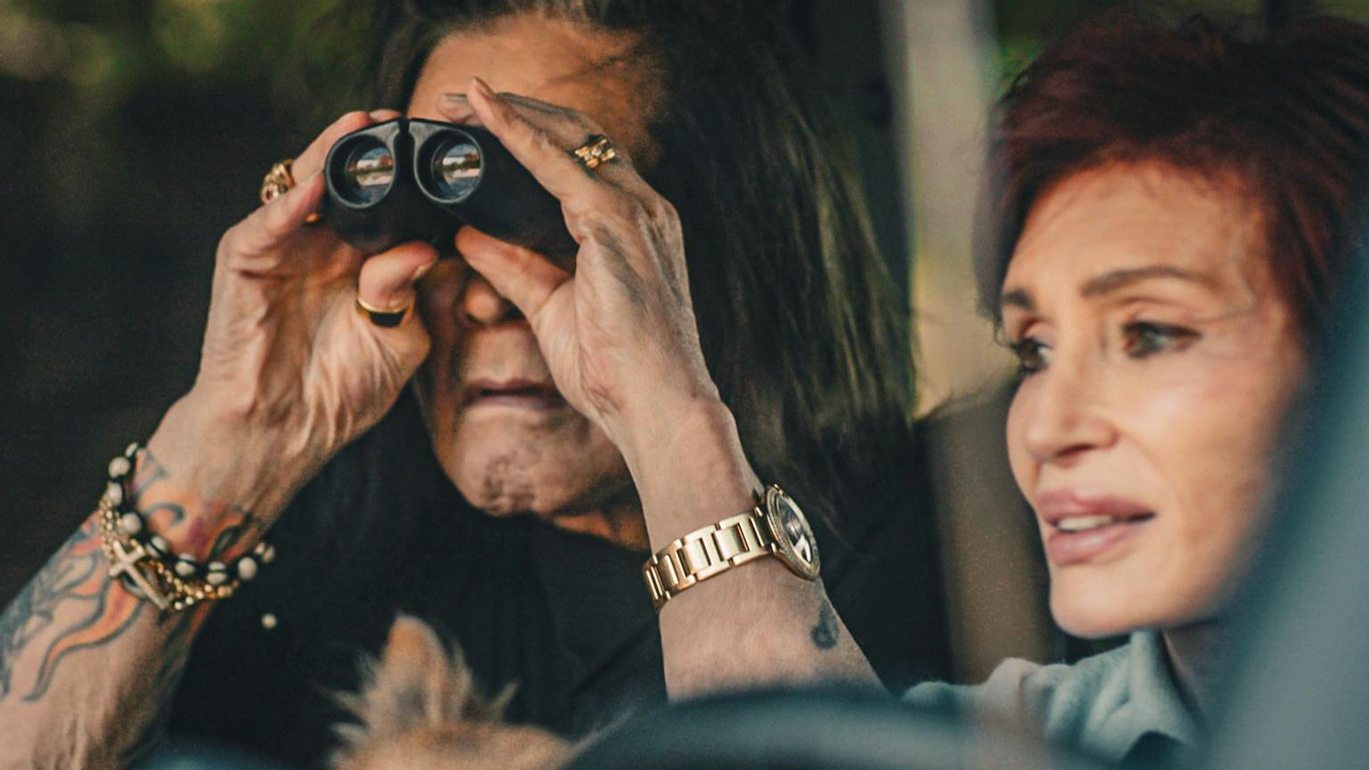 Passenger Ozzy Osbourne looks out of the front window of a vehicle through binoculars, as Sharon, sat next to him, drives