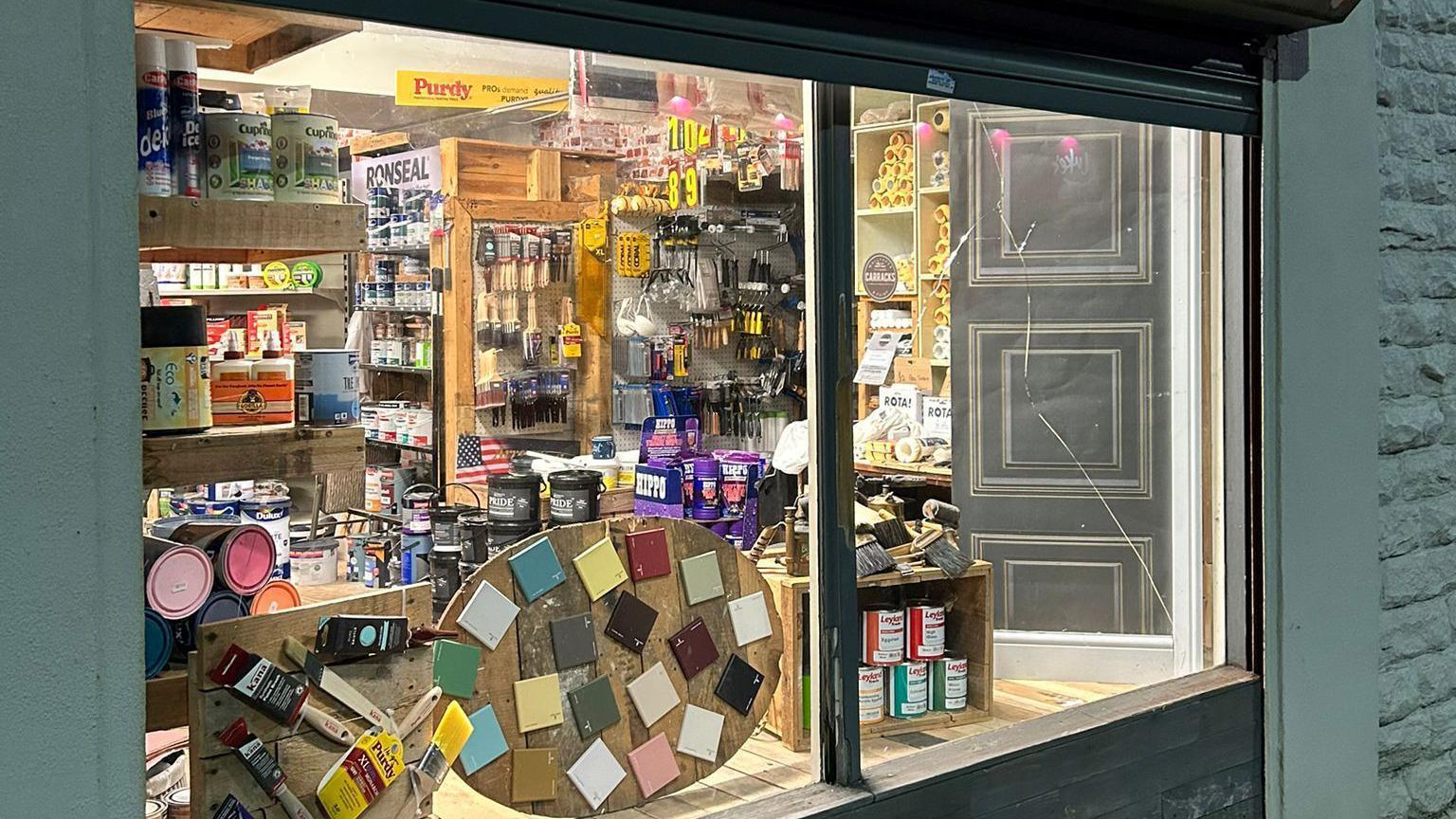 The front window display of a shop. Inside the window, shelves are packed with various decorating supplies including paint tins, brushes and rollers.