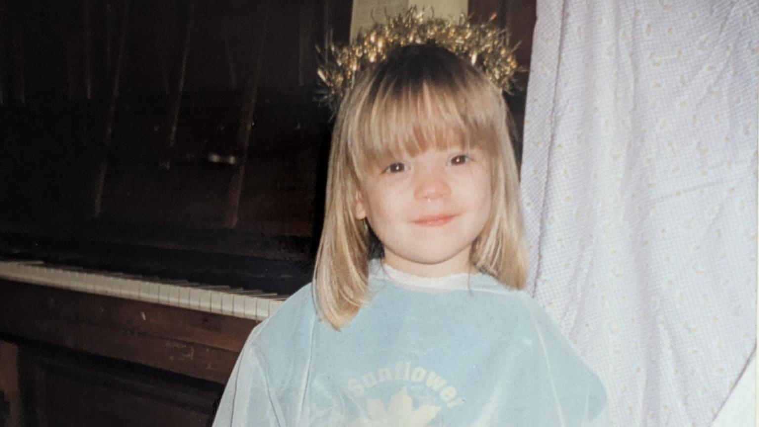 Amy as a young child wearing a light blue top and a tinsel crown