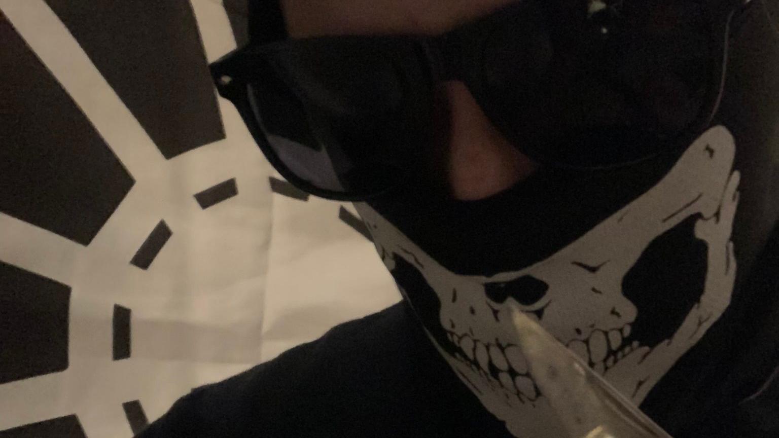 A man alleged to be Alfie Coleman wearing a balaclava with a skull on it and sunglasses while holding a knife. A black flag with a white spider's web is pinned to a wall behind him.