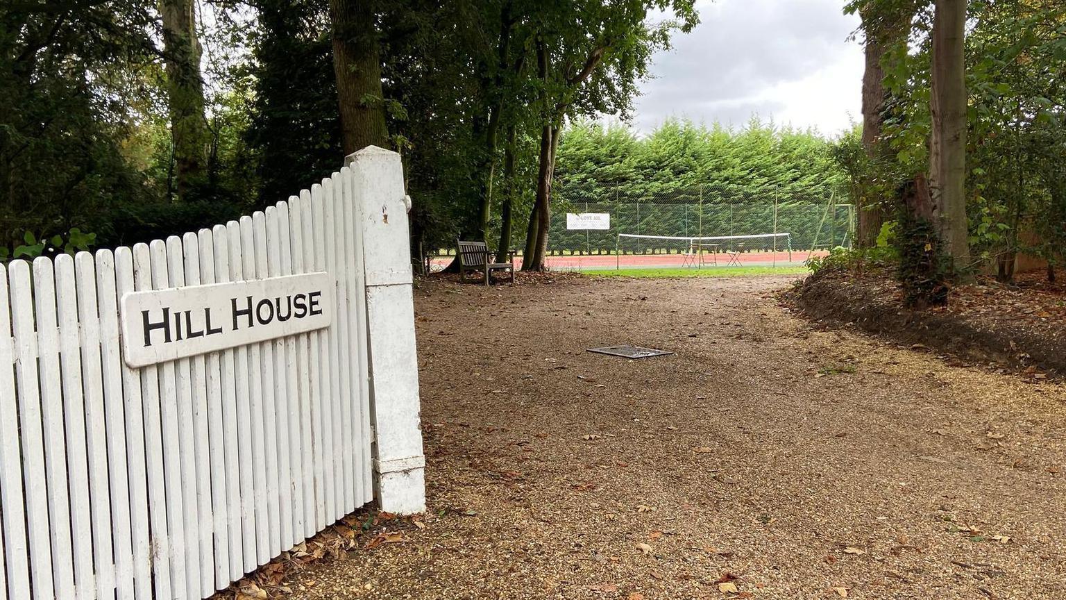 An entrance to the Hill House property. White fences line the entrance with the name of the house written on the fence. A tennis court can be seen in the distance and trees line either side of the driveway