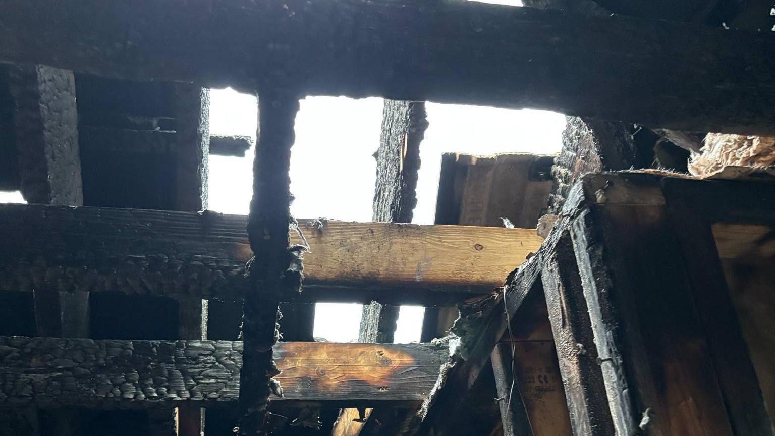 The gutted family home. All of the beams are charred and their is black everywhere. Burned items can be seen on the floor.