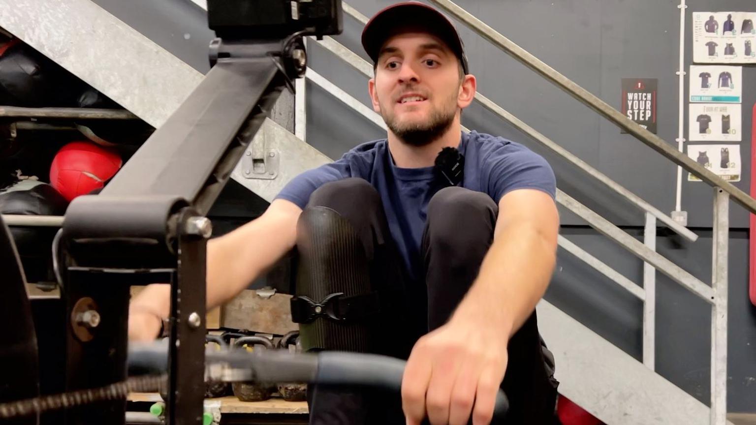 A man in blue T-shirt and a cap on rowing machine in a gym.