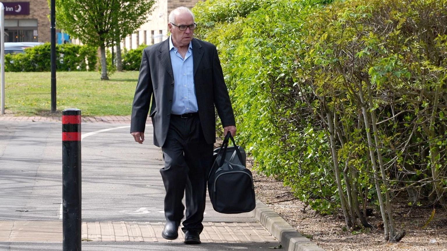 Kevin Miller is wearing a grey or navy blue suit while walking on a pavement and carrying a dark holdall in his left hand.