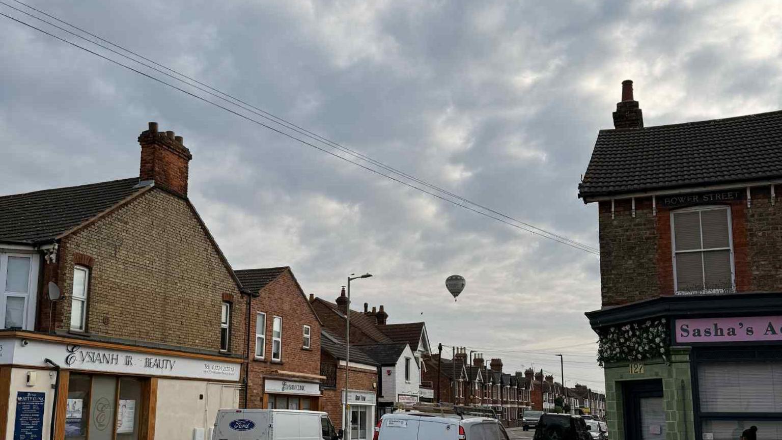 A hot air balloon over the Castle Road area of Bedford, showing rows of buildings shops, brick terraced houses, telegraph cables, wires, lamp posts, in the sky, that is grey. There are vehicles on the road.