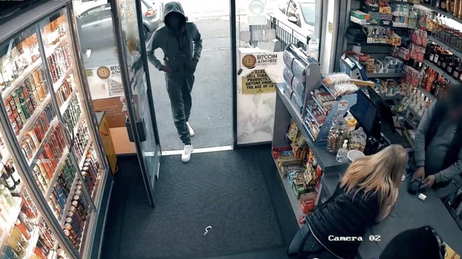 A CCTV image of a man walking into a convenience store as a woman leans over a counter while a shop work enters details into a card machine. 