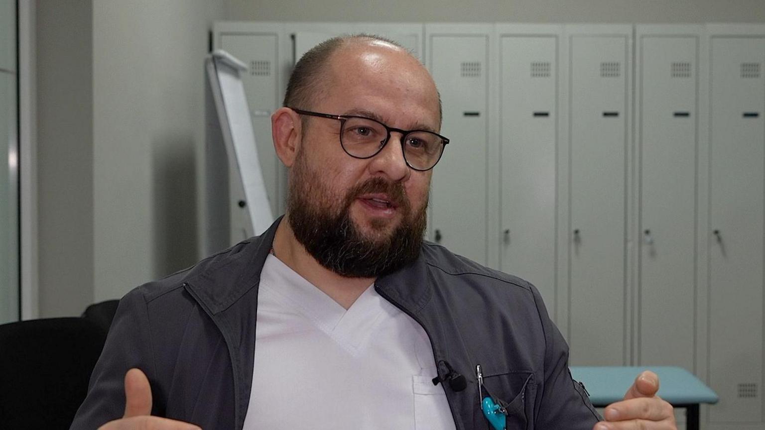 Balding man with beard in glasses sits in a room with lockers in the background