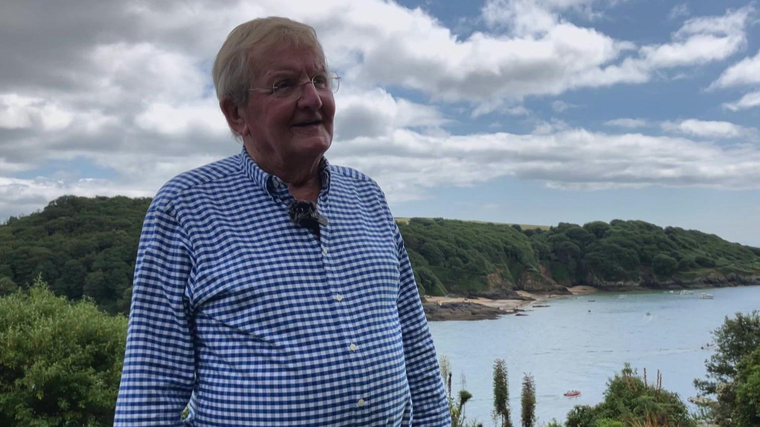 Simon Henn looking to the right. He is wearing a white and blue checked shirt and to his right is a sea view. There is a sandy beach below and the blue sea. Around the