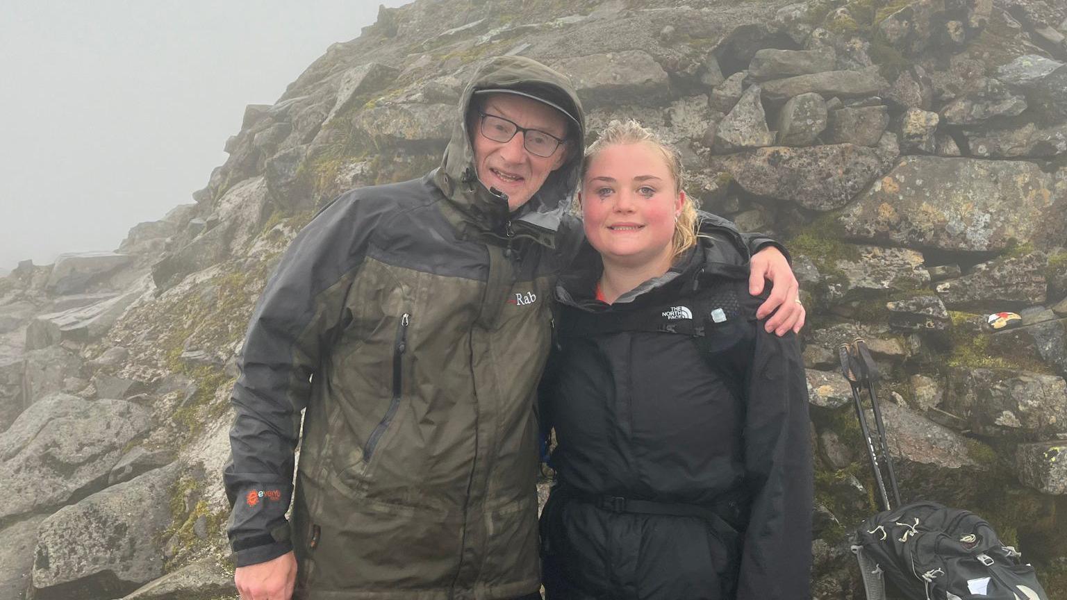 A man and his daughter stood on the top of a rocky mountain. it is foggy. There are rocks around them.