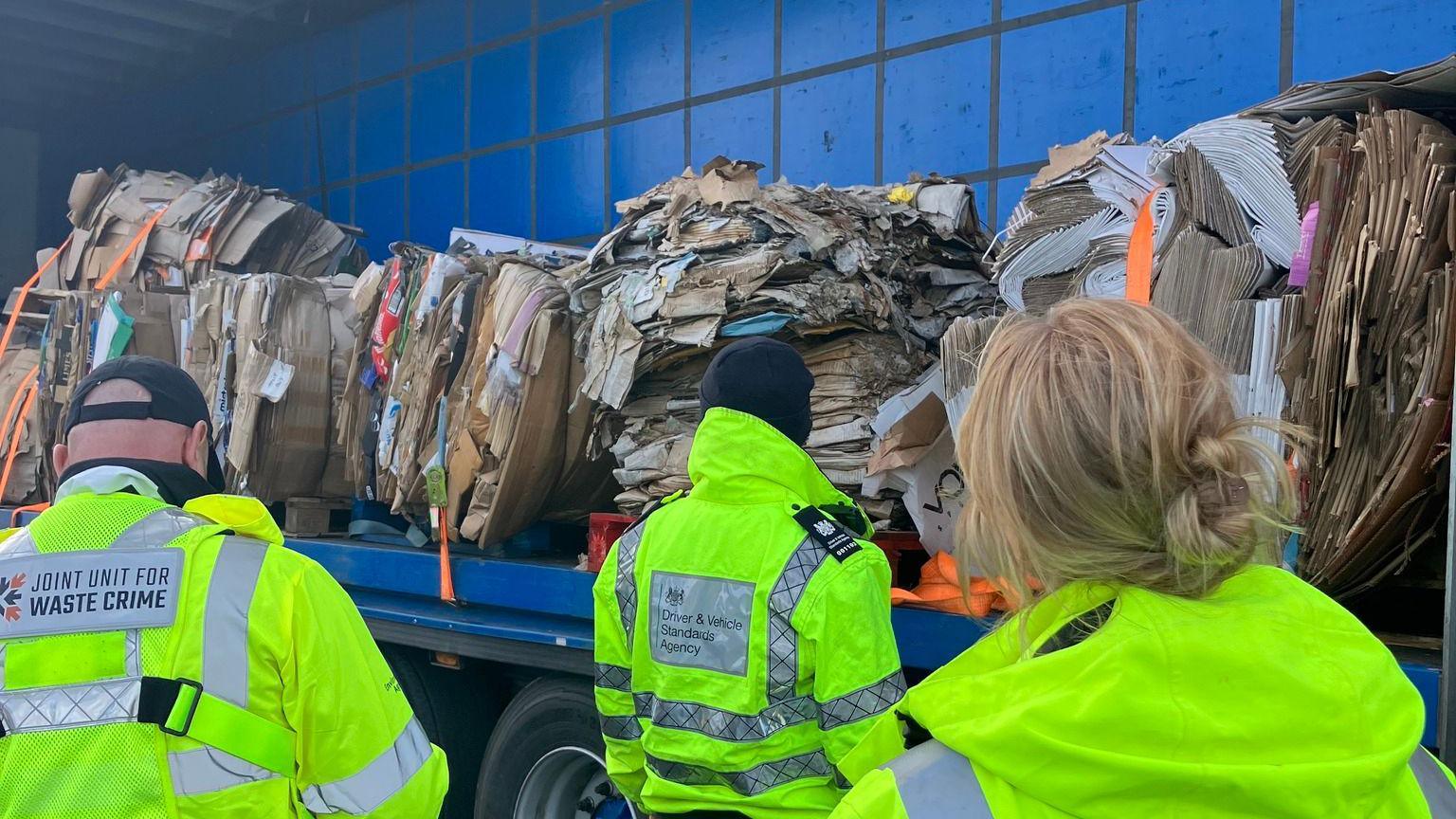 A blue lorry is opened on one side which shows piles of paper and cardboard bundled. There people wearing hi-vis jackets are looking at the lorry.