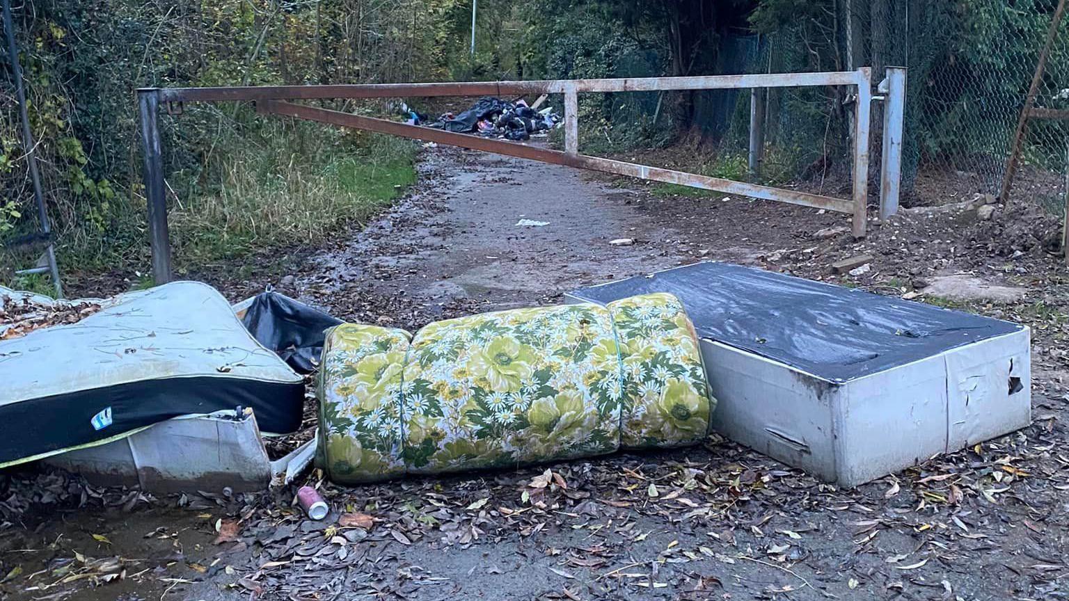 Dumped mattresses and a divan bed-base in front of a shut metal gate in front of a path through woodland. The mattress and divan base are white and a rolled mattress with green, yellow and white flowers is in the centre of the picture. In the far background a mound of black bags can be seen.