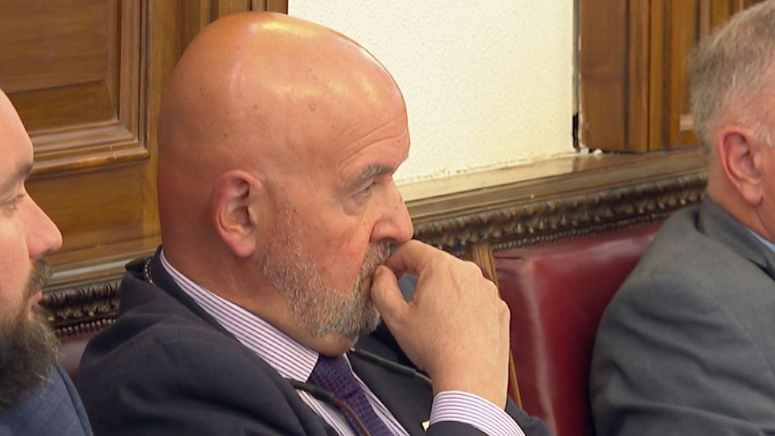 Deputy Jonathan Le Tocq - A bald man with a short beard with his hand on his mouth. He's looking away from the camera so we see his side profile.