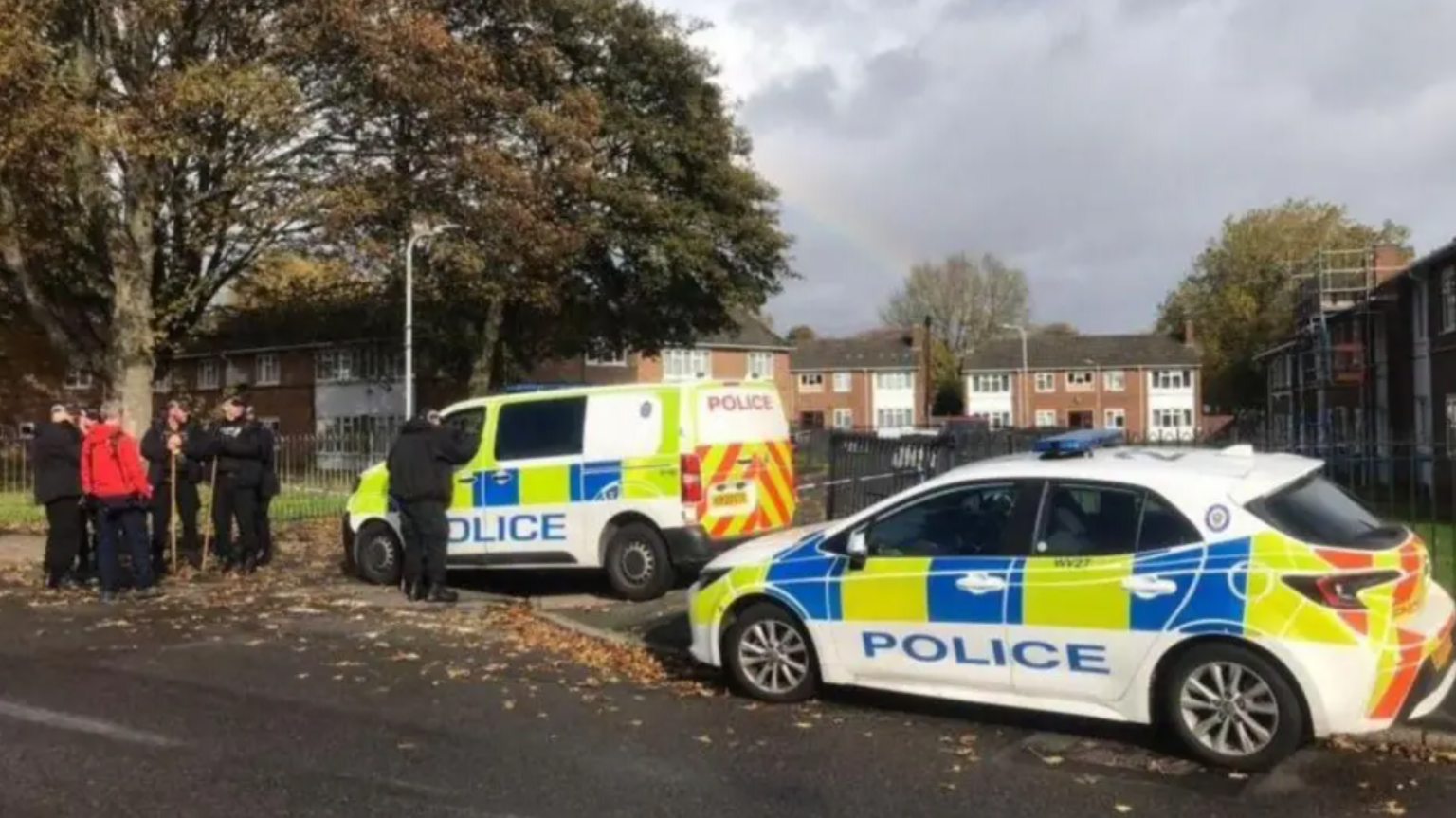 A police car and a police van block access to a residential side street. Several police officers wearing all black, some holding long sticks, are standing on the left with a person wearing a red top. They are underneath a large tree. Terraced houses can be seen in the distance.