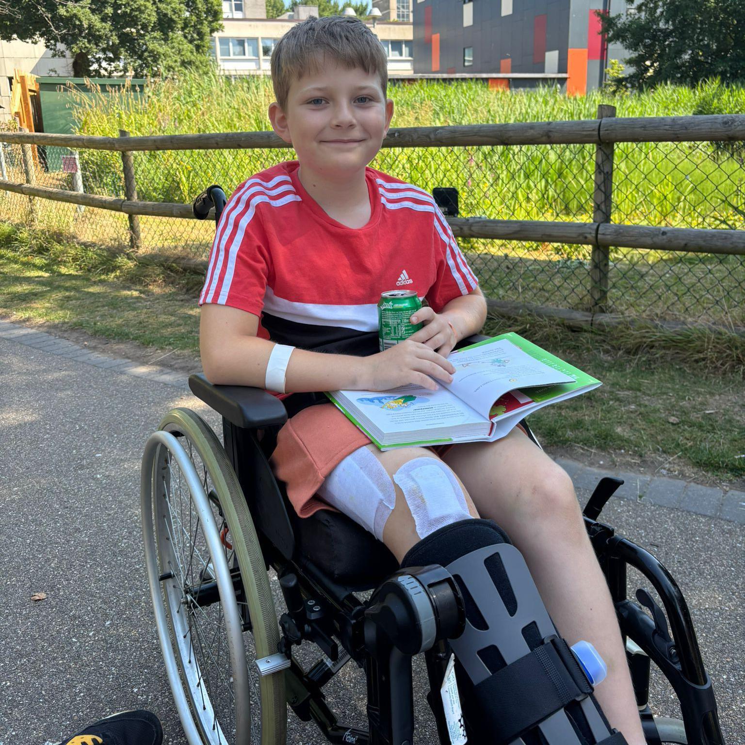 Theo, who has short light brown hair, is wearing a red t-shirt and sat in a wheelchair with a drink can and a book open on his lap. A fence and a patch of long grass can be seen behind him.